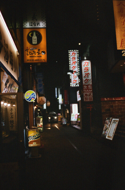 A narrow, dimly lit street at night in Japan, lined with brightly glowing vertical and horizontal signs in Japanese. The atmosphere is urban and quiet, with no people visible.