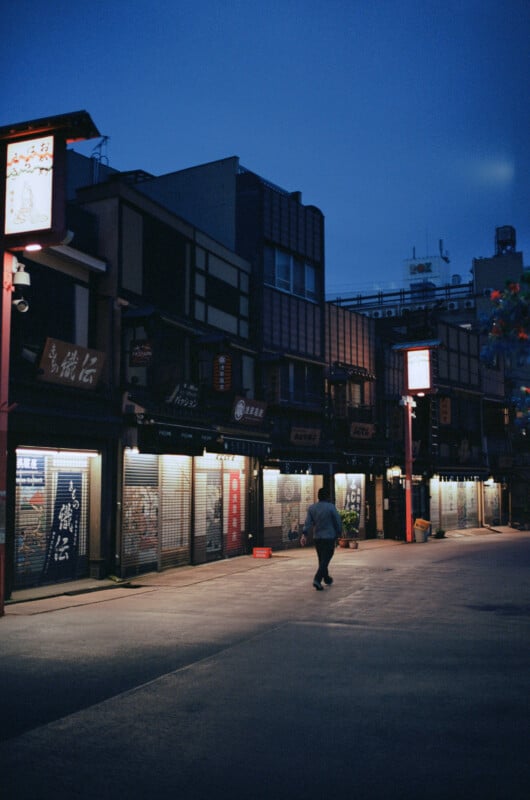A person walks alone down a quiet street lined with closed shops and illuminated signs at dusk, creating a peaceful, atmospheric scene in an urban area.