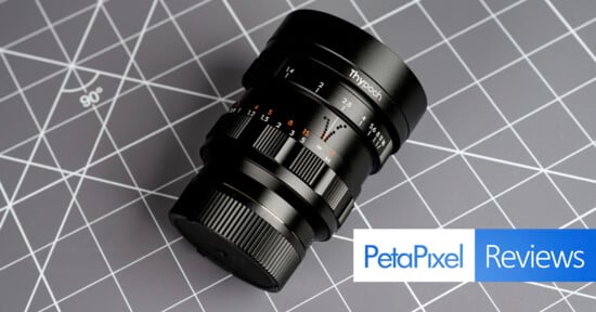 A black camera lens with focus markings rests on a gray cutting mat with white grid lines. The Petapixel logo and the word "Reviews" are displayed in the lower right corner.