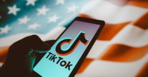 A hand holding a smartphone displaying the TikTok logo, with a blurred United States flag in the background.