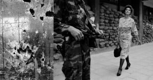 A child looks through a shattered glass window on the left, while a soldier with a gun patrols and a woman walks confidently past sandbags on the right. The scene appears tense and contrasts innocence, authority, and resilience.