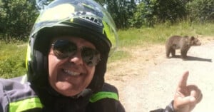 A person wearing a motorcycle helmet and sunglasses smiles for a selfie while pointing at a brown bear standing on a dirt path in a sunny, wooded area.