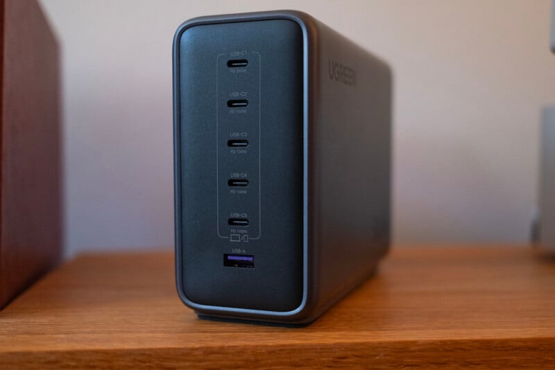 A black charging station with five visible USB ports, four USB-C ports labeled with power outputs and one USB-A port, sits on a wooden shelf against a plain background.