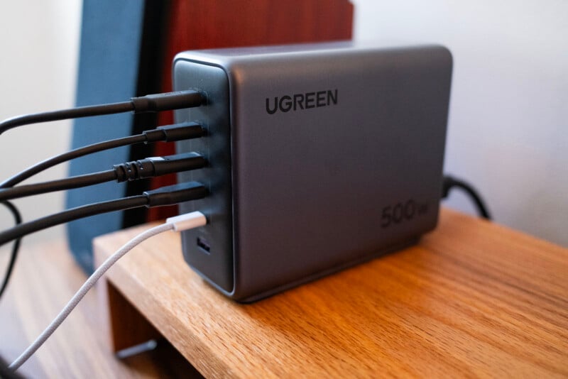 A UGREEN 500W charging hub sits on a wooden shelf, with multiple black and one white cable plugged into its ports. The device is in use in a home or office setting.