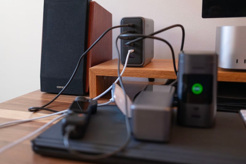 A close-up of a cluttered desk with various electronics, including a speaker, multiple charging devices, cables, and a laptop, all placed on a wooden surface and a small shelf.