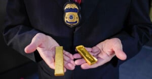A person in a dark suit with a Homeland Security Investigations badge holds two gold bars, one in each hand.