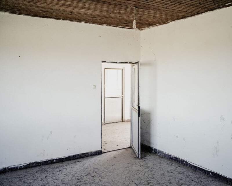 An empty room with white walls, a wooden ceiling, and a single light bulb hanging from the center. A partially open door leads to a sunlit, dust-covered area beyond. The floor is bare and dusty.