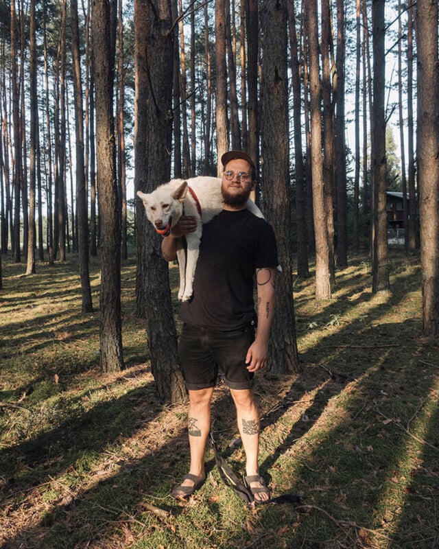 A man with a beard and glasses, dressed in black shorts and a t-shirt, stands in a sunlit pine forest with a white dog draped over his shoulders. Both look at the camera. A wooden cabin is visible in the background.