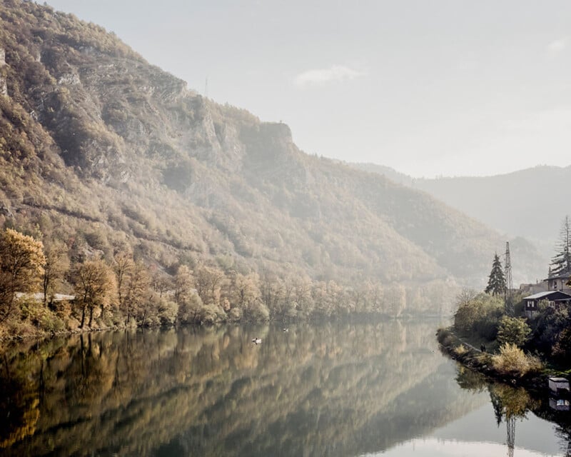 A calm river reflects tree-covered hills and mountains under a hazy sky, with some houses and greenery on the right bank.