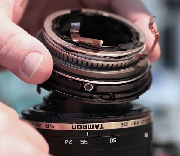 A close-up of a person's hand repairing or assembling the upper part of a Tamron camera lens, showing detailed internal components and circuits.
