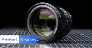 A camera lens rests on a reflective, grid-patterned surface with the "PetaPixel Reviews" logo in the lower left corner. The lens is angled to show the front glass and detailed markings.