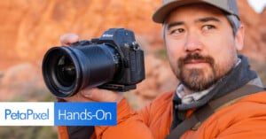 A person wearing an orange jacket and a cap holds a Nikon camera, aiming it at something off-camera. In the foreground, there's text overlay saying "PetaPixel Hands-On." The background shows a blurred, rocky landscape.