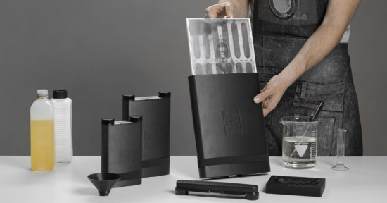 A person wearing a dark apron prepares film development chemicals and equipment on a white table, including black containers, bottles, a beaker, a measuring cylinder, a funnel, and a sheet of film being inserted into a black tank.